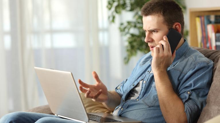 Schmuckbild: Wütender Mann spricht zu Hause am Telefon. Er hat einen Laptop auf dem Schoß.