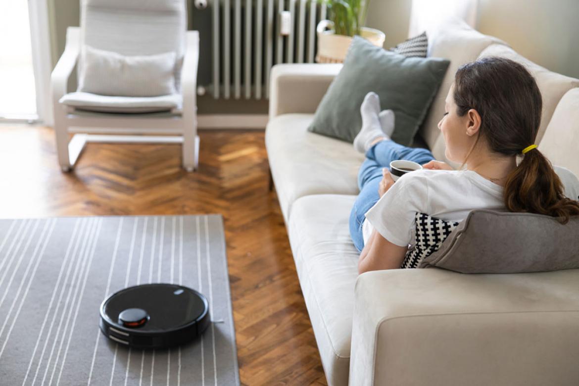 Frau mit Kaffee auf Sofa, davor ein Saugroboter