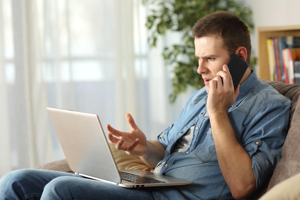 Schmuckbild: Wütender Mann spricht zu Hause am Telefon. Er hat einen Laptop auf dem Schoß.