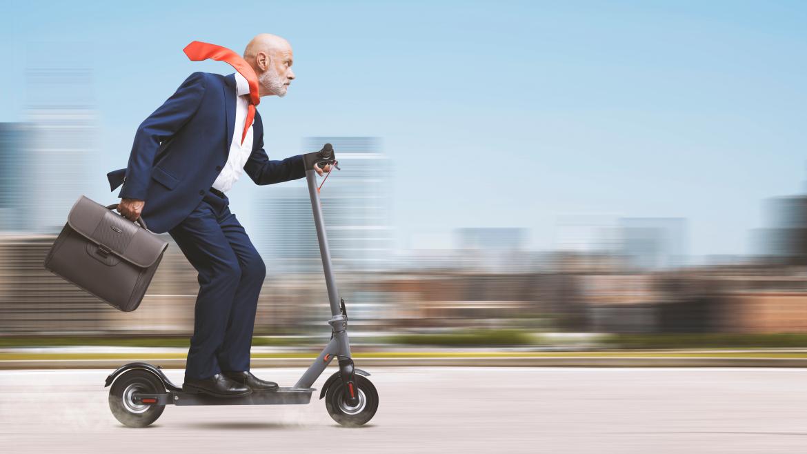 Mann im Anzug fährt auf E-Roller zur Arbeit. Die Krawatte weht im Wind.