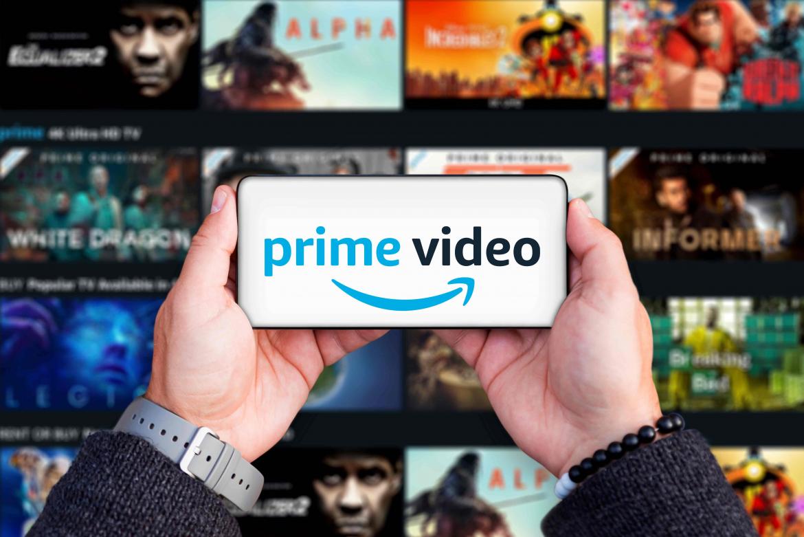 Händ ehalten Handy auf dem der Schriftzug prime Video zu sehen ist. Im Hintergrund sieht man den die Prime Fimauswahl auf einem großen Fernseher 